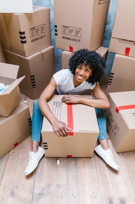 Two women standing inside a room surrounded by packed cardboard boxes, some sealed with red tape, indicating a home relocation process. One woman, with long brown hair and wearing a plaid shirt over a