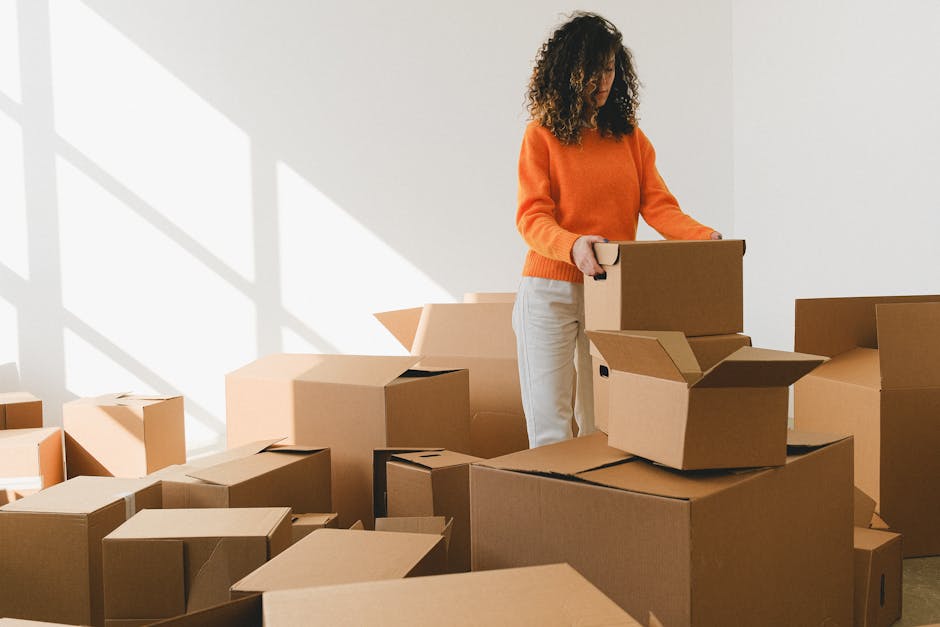 A woman with curly hair dressed in an orange sweater and light-coloured trousers is inside a well-lit room, surrounded by numerous brown cardboard boxes of various sizes, some sealed with packing tape and others open with flaps. She is in the process of packing or preparing to move, holding a small box while standing amidst the boxes that are stacked or placed on the floor. The sunlight streams through a large window, casting shadows on the plain white wall behind her. The scene depicts the packing stage of a home relocation, with the boxes arranged for transport, supported by a moving service such as Man with Van Brockley. The environment suggests a typical interior of a Victorian Brockley home engaged in furniture transport and packing. No furniture or appliances are visible, but the focus on packing materials and the spatial arrangement emphasizes the logistics involved in house removals.