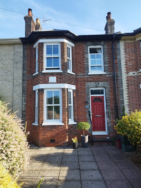 A two-storey Victorian-style brick house with a red front door, located in Brockley, London. The building features white window frames and a bay window on the ground floor, with a small white plaque mounted on the brick wall above. The pathway leading to the entrance is paved with concrete slabs, flanked by well-maintained shrubs and potted plants, including a small tree in a black pot near the door. The sky is clear and blue, with chimney stacks visible on the roof. The image captures the exterior of the property, illustrating its traditional architectural details. As part of house removals and furniture transport, [COMPANY_NAME], such as Man with Van Brockley, would coordinate the logistics of packing and moving items securely in such residential settings, ensuring minimal risk of damage during the loading process.