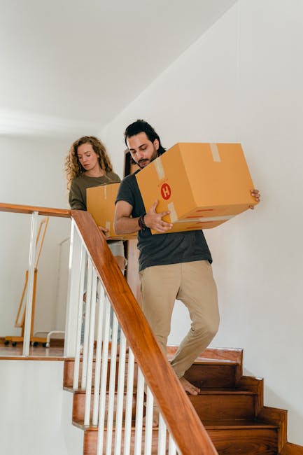 A woman with curly hair dressed in an orange sweater and light-coloured trousers is inside a well-lit room, surrounded by numerous brown cardboard boxes of various sizes, some sealed with packing tape and others open with flaps. She is in the process of packing or preparing to move, holding a small box while standing amidst the boxes that are stacked or placed on the floor. The sunlight streams through a large window, casting shadows on the plain white wall behind her. The scene depicts the packing stage of a home relocation, with the boxes arranged for transport, supported by a moving service such as Man with Van Brockley. The environment suggests a typical interior of a Victorian Brockley home engaged in furniture transport and packing. No furniture or appliances are visible, but the focus on packing materials and the spatial arrangement emphasizes the logistics involved in house removals.