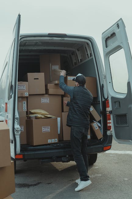 A young man is standing on a residential street next to a white van with its side door open, engaged in the packing and loading process for home relocation. He is holding a cardboard box and is surrou