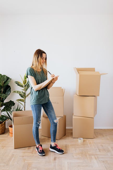 A young woman in casual clothing, wearing a green t-shirt and ripped blue jeans, standing in a well-lit indoor space next to several stacked cardboard boxes, with one box open. She is holding a pen and a clipboard, appearing to be taking notes during a home relocation or packing process. To her left, there is a potted plant with large green leaves placed on the floor. The background features a plain white wall, and the floor is made of light-colored wood. The scene depicts the packing stage of a house move, with boxes prepared for transport, aligning with services offered by Man with Van Brockley for furniture transport and move organization.