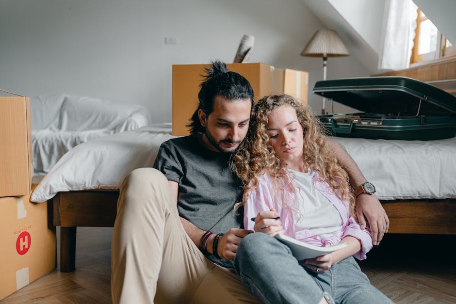 A man with dark hair tied back and a woman with curly blonde hair are sitting on a wooden floor inside a room, leaning against a bed with white bedding, during a home relocation. The man is wearing a dark grey t-shirt and beige trousers, holding a smartphone, while the woman is dressed in a white top with a light pink cardigan, holding a clipboard and pen, appearing to be planning or organizing. In the background, there are several cardboard boxes, some with packing tape, and a green hard-shell suitcase open on the bed, indicating packing and moving preparations. The room has natural light coming through a window with sheer curtains, and a bedside lamp on a wooden nightstand is visible. Man with Van Brockley often assists with furniture transport and packing services for house removals, supporting clients during the loading process of their belongings onto a van, which may occur nearby outside the property. The environment suggests an active moving day with organized packing and logistical planning.