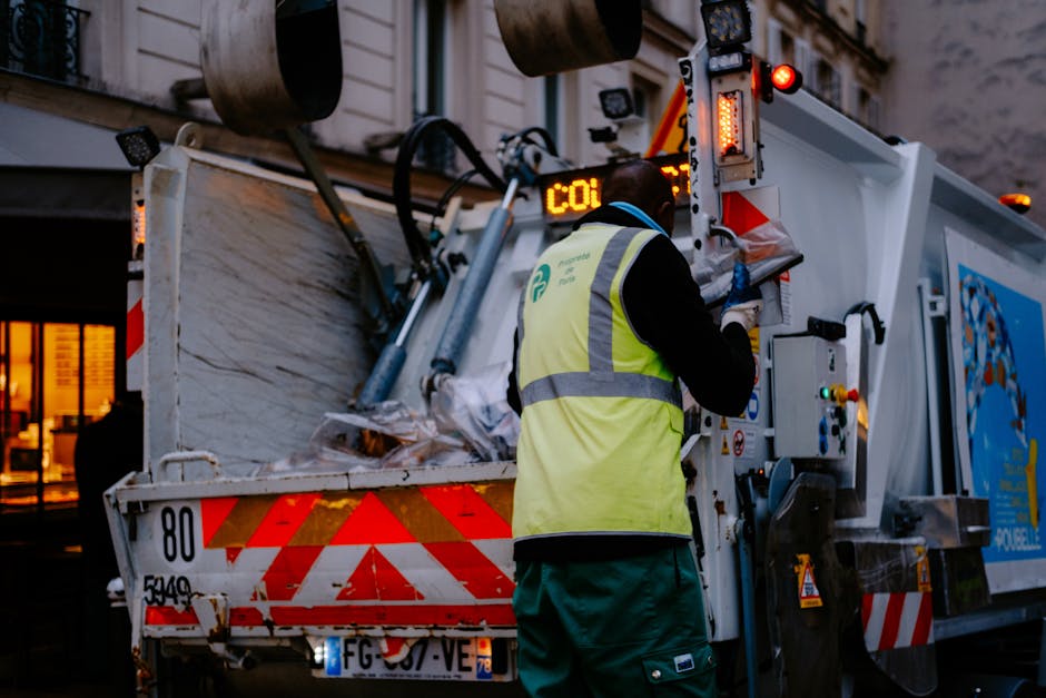 A waste collection worker in a high-visibility yellow vest with reflective stripes is operating a rubbish truck in an urban area during late evening or early night, as indicated by the dim lighting. The worker, facing away from the camera, is engaged in a loading process, possibly collecting bulky waste or debris as part of a house removal or clean-up service. The rubbish truck features various mechanical components, including hydraulic arms, control panels, and warning lights, with a blue graphic and logo partially visible on its side. The rear of the truck is open, revealing a large compartment filled with assorted waste materials such as cardboard boxes, plastic packaging, and wooden items, some of which are wrapped in protective materials like fabric or plastic sheeting. The scene focuses on the logistical aspect of waste removal associated with home relocation or moving services, highlighting the importance of proper clearance procedures. Man with Van Brockley occasionally undertakes such removal operations, supporting clients with disposal and transport of unwanted items efficiently.