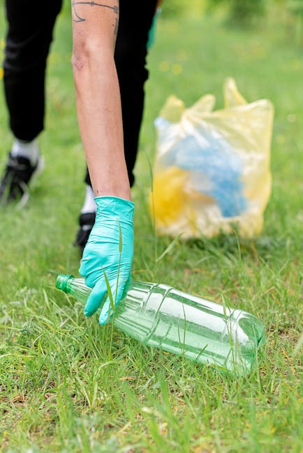Close-up of a person wearing a blue glove picking up an empty, transparent plastic bottle from the grass, with a yellow and blue rubbish bag in the background. The scene is outdoors on a grassy area, possibly during a cleanup or waste collection activity. The person's arm and hand are visible, with the focus on the action of collecting recyclable material, reflecting environmentally responsible waste disposal. This image relates to house removals and clearing rubbish as part of a home relocation or clearance service offered by Man with Van Brockley, highlighting the importance of proper waste management during moving or decluttering processes.