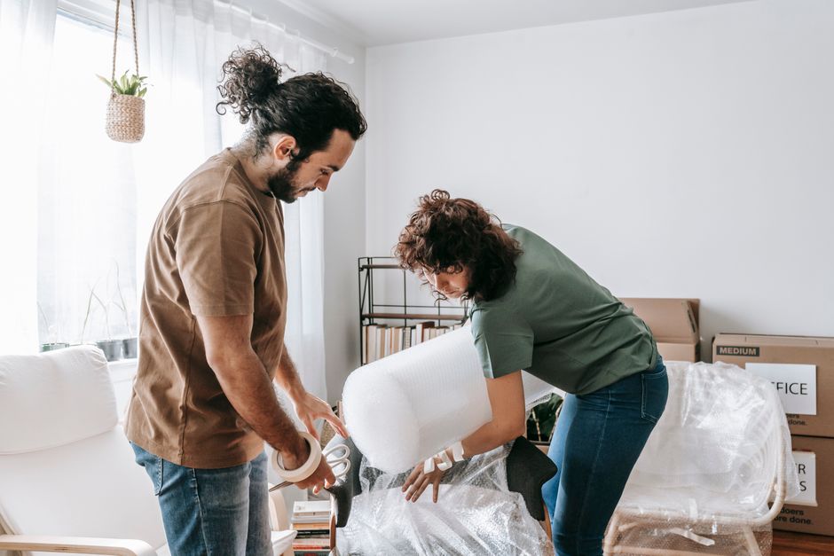Two movers from Man with Van Brockley are inside a well-lit room preparing to load a large piece of furniture wrapped in white protective padding onto a moving van. The male mover, wearing a brown t-s
