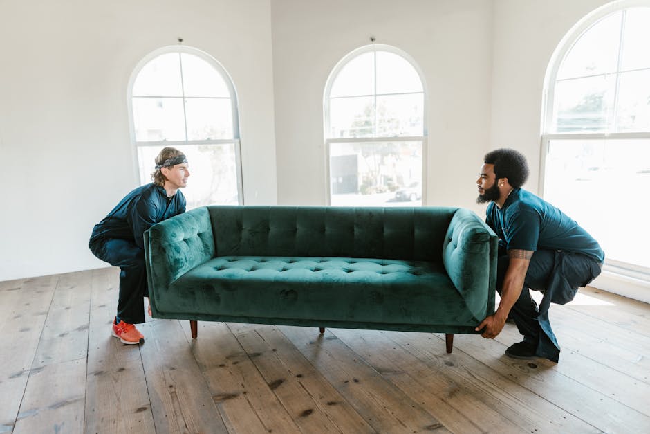 Two movers from Man with Van Brockley are inside a well-lit room preparing to load a large piece of furniture wrapped in white protective padding onto a moving van. The male mover, wearing a brown t-s