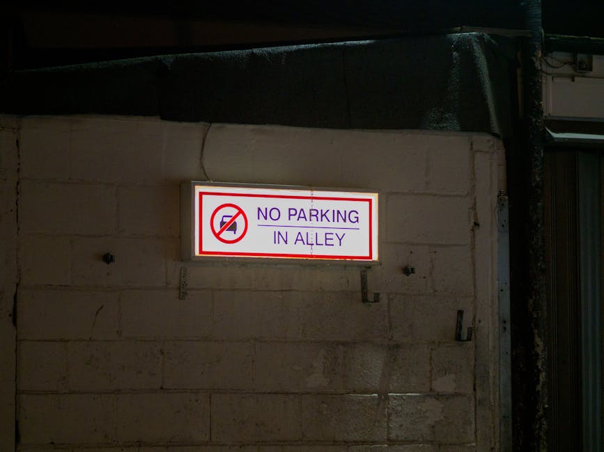 A rectangular illuminated sign mounted on a beige brick wall, displaying the message 'NO PARKING IN ALLEY' in purple text, with a red circle and slash over a black car icon, indicating parking restrictions. The sign is attached near an entrance or exit area, with partial view of a metal door frame to the right. The lighting suggests it is nighttime or in a dimly lit environment, with the sign providing clear visibility for drivers and pedestrians. The setting appears to be an alley or rear access point, relevant to home relocation or furniture transport services provided by Man with Van Brockley, which may operate in areas requiring compliance with parking regulations during removals or moving activities.