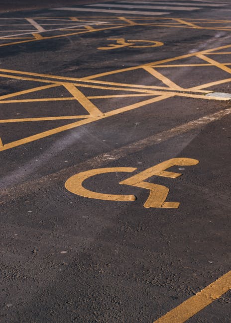 Close-up view of a parking lot surface featuring multiple yellow painted parking space markings, including designated spaces for disabled parking with the universal wheelchair symbol. The asphalt ground shows some wear and tire marks, with the yellow lines and symbols clearly visible and freshly painted. The image emphasizes the spatial arrangement of parking bays, highlighting accessibility zones that may be relevant for vehicle parking during house removals or furniture transport. Such parking lot markings are commonly seen outside residential or commercial properties in Brockley, where Man with Van Brockley provides removals and moving services, ensuring proper parking compliance for loading and unloading during home relocation tasks.