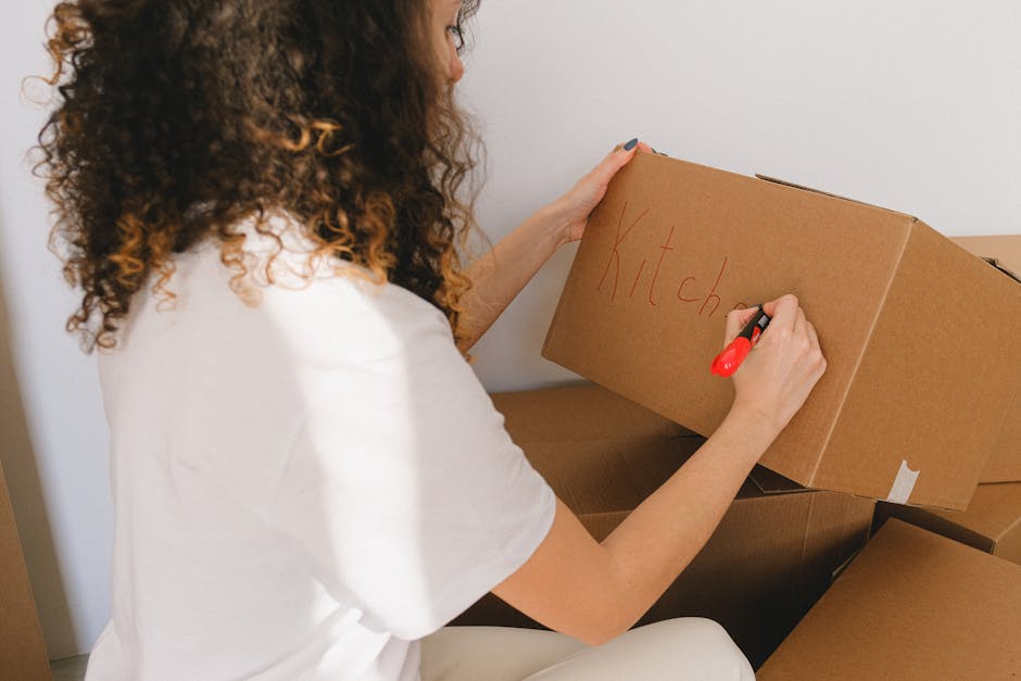 A young woman with curly black hair, dressed in a white top, blue jeans, and white sneakers, sits on the wooden floor inside a room surrounded by numerous cardboard moving boxes of various sizes. Some boxes are sealed with red packing tape, while others are open or partially collapsed. The boxes are stacked against the blue and white textured wall, creating a cluttered atmosphere typical of a home in the process of packing for a move. The woman appears happy and relaxed, resting her arms on a sealed cardboard box in front of her. Nearby, a small white piece of packing material or tissue is on the floor. The scene depicts the final stages of packing and preparation for a home relocation, with lighting that highlights the details of the boxes and the woman's cheerful expression. This visual aligns with the moving and packing services offered by Man with Van Brockley, supporting the page's focus on local removals tips in the Telegraph Hill Park area.