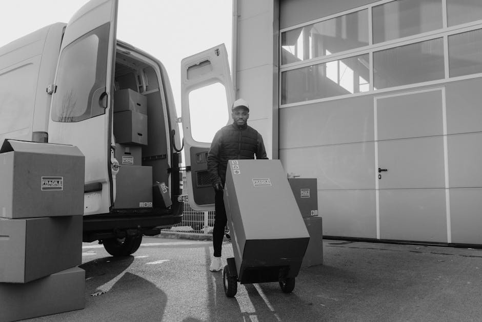 A man wearing a dark jacket and cap is standing outside a delivery van with its side door open, unloading large cardboard boxes and plastic-wrapped items onto the pavement near a modern building with glass windows and metal panels. Several cardboard boxes are arranged on the ground, some labeled with fragile stickers, and a hand truck is being used to transport one of the boxes. The scene takes place in natural daylight, capturing a home relocation or furniture transport process as part of house removals services by Man with Van Brockley, with the loading and packing materials positioned close to the building's entrance for efficient moving logistics.