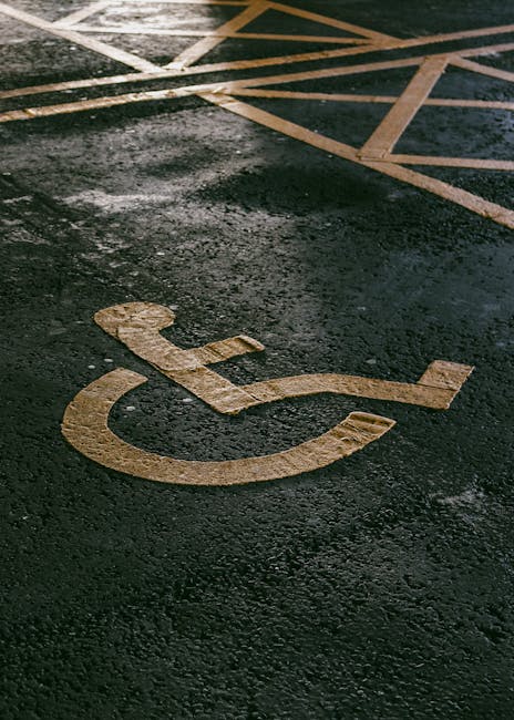 A designated accessible parking space with a white painted wheelchair symbol on a dark asphalt surface, partially wet with reflections and slight texture visible. In the background, there are yellow and white striped markings on the ground indicating a no-parking or restricted area, with some paving stones visible along the edge. The scene is outdoors and well-lit, likely during daylight hours. This image relates to the logistics of home relocation, demonstrating parking considerations for furniture transport and moving services. Occasionally, Man with Van Brockley, providing removals in SE4, utilises such spaces for efficient loading and unloading during residential moves, including unloading packed boxes, furniture, and appliances from a moving van. The environment is typical of a driveway or parking area adjacent to a residential property, emphasizing the importance of parking arrangements in planning a house move, with clear visual cues for accessibility and moving logistics.