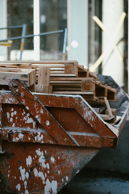 A close-up of a rusted metal boat used as a makeshift platform for transporting wooden pallets and packaging materials during a house relocation. The boat is loaded with stacked wooden pallets, some with slats and others with thicker beams, secured for transport. The pallets are surrounded by remnants of white paint splatters on the boat's exterior, indicating it has been repurposed outside. In the background, part of a modern glass building and a white weather vane are visible, suggesting an urban or suburban setting. The scene captures a loading process likely managed by Man with Van Brockley, highlighting the careful handling of furniture and packing materials involved in removals and furniture transport services.