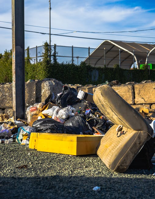 An outdoor scene showing a large pile of black and clear plastic rubbish bags, some torn open, alongside discarded items such as a worn, dirty car tire leaning against the pile, and a yellow plastic crate on a gravel surface. In the background, there is a low stone wall, a metal utility pole, overhead electrical wires, and a partially visible green hedge. The sky is partly cloudy with blue patches overhead. The image appears to depict waste or debris which may be associated with bulky collection or removal processes, relevant to house removals and packing logistics. Man with Van Brockley services focus on efficient removal and clearance, often involving careful handling of furniture, boxes, and appliances during home relocation. The scene highlights the importance of proper disposal and planning in moving operations.