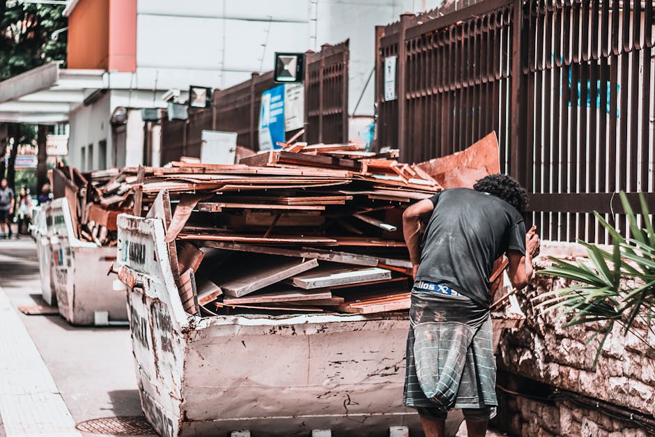 A close-up of a rusted metal boat used as a makeshift platform for transporting wooden pallets and packaging materials during a house relocation. The boat is loaded with stacked wooden pallets, some with slats and others with thicker beams, secured for transport. The pallets are surrounded by remnants of white paint splatters on the boat's exterior, indicating it has been repurposed outside. In the background, part of a modern glass building and a white weather vane are visible, suggesting an urban or suburban setting. The scene captures a loading process likely managed by Man with Van Brockley, highlighting the careful handling of furniture and packing materials involved in removals and furniture transport services.