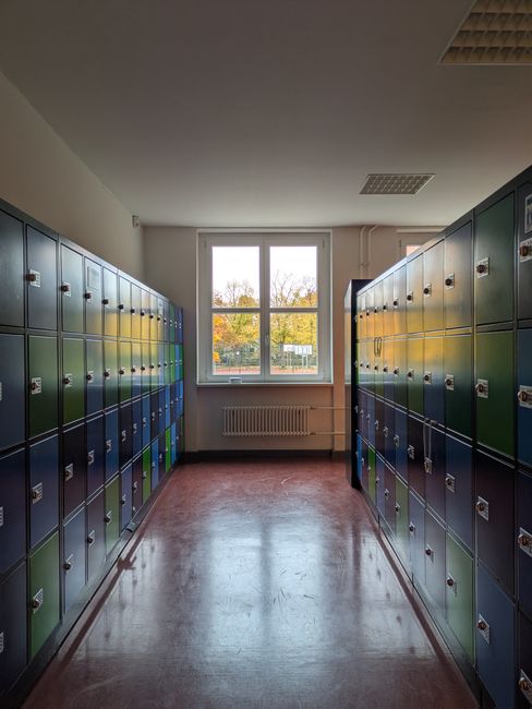 The image depicts a narrow indoor corridor with a row of green and blue metal lockers on both sides, each locker equipped with a small ventilation slot and a lock. The corridor is illuminated by natural light coming through a large window at the far end of the space, revealing trees with autumn foliage outside. The floor appears to be made of polished linoleum or vinyl, reflecting the light from the window. Below the window, there is a white radiator attached to the wall. The ceiling is fitted with recessed lighting panels. The environment suggests a school or communal locker area, which may be relevant in the context of a house removal or relocation involving storage or packing solutions. Man with Van Brockley, a removal service company, might utilize such spaces during furniture transport or storage arrangements for clients undertaking home relocations, including student moving or storage between lets.