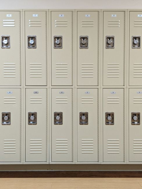 A row of beige metal lockers with small, built-in combination locks and ventilation slits, arranged in two tiers on a wall inside a building corridor with a wooden floor. The lockers are numbered with small blue labels, indicating their individual storage compartments. The lighting is neutral, providing clear visibility of the lockers' surface and details. This image is related to home relocation services, such as packing and moving, as offered by Man with Van Brockley, especially in the context of storing belongings for students between lets in Brockley. The lockers symbolize storage solutions used during furniture transport or temporary storage phases in house removals and moving processes.
