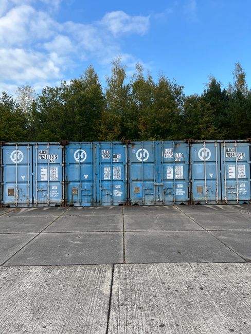The image depicts a narrow indoor corridor with a row of green and blue metal lockers on both sides, each locker equipped with a small ventilation slot and a lock. The corridor is illuminated by natural light coming through a large window at the far end of the space, revealing trees with autumn foliage outside. The floor appears to be made of polished linoleum or vinyl, reflecting the light from the window. Below the window, there is a white radiator attached to the wall. The ceiling is fitted with recessed lighting panels. The environment suggests a school or communal locker area, which may be relevant in the context of a house removal or relocation involving storage or packing solutions. Man with Van Brockley, a removal service company, might utilize such spaces during furniture transport or storage arrangements for clients undertaking home relocations, including student moving or storage between lets.