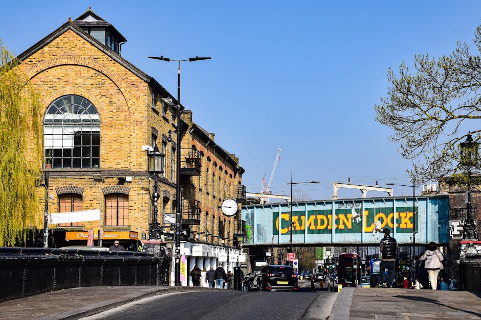 A street scene in Brockley featuring a multi-story brick building with large arched and rectangular windows, some with visible windowpanes, under a clear blue sky. On the street level, there are pedestrians walking along the pavement, some near storefronts including one with an orange sign displaying 'Orange Juice.' In the foreground, a black van is partially visible, parked adjacent to the pavement. Overhead, a large railway bridge painted with a weathered green surface bears the bold yellow text 'CADDIN LOCK.' The bridge spans the width of the street, with the railway tracks and overhead electrical lines above. To the right, a leafless tree extends its branches into the sky, and street lamps and a clock are mounted on poles along the road, contributing to the urban environment. The scene depicts a busy city street in Brockley with active pedestrian movement, vehicle parking, and transportation infrastructure activity, consistent with typical house removals and logistics surrounding packing and furniture transport, as performed by service providers such as Man with Van Brockley.
