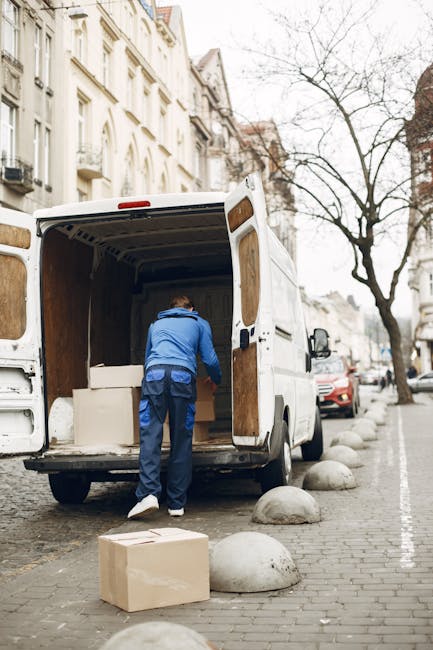 A man dressed in a blue jacket and dark trousers is seen loading a cardboard box into the open rear of a white moving van parked on a cobbled street. The van's interior reveals several large wrapped furniture items, including a white piece that appears to be a small wardrobe or storage unit, supported by the van's wooden side paneling. Outside, on the street, there are more cardboard boxes and grey concrete bollards separating the road from the pavement. In the background, residential buildings with classic architectural details and white facades line the street, while a leafless tree stands nearby under an overcast sky, indicating the scene takes place during a typical home relocation or furniture transport process. This visual captures a professional removals service, such as Man with Van Brockley, involved in packing and loading belongings for a house move, emphasizing careful handling of household items during a furniture transport in an urban area.